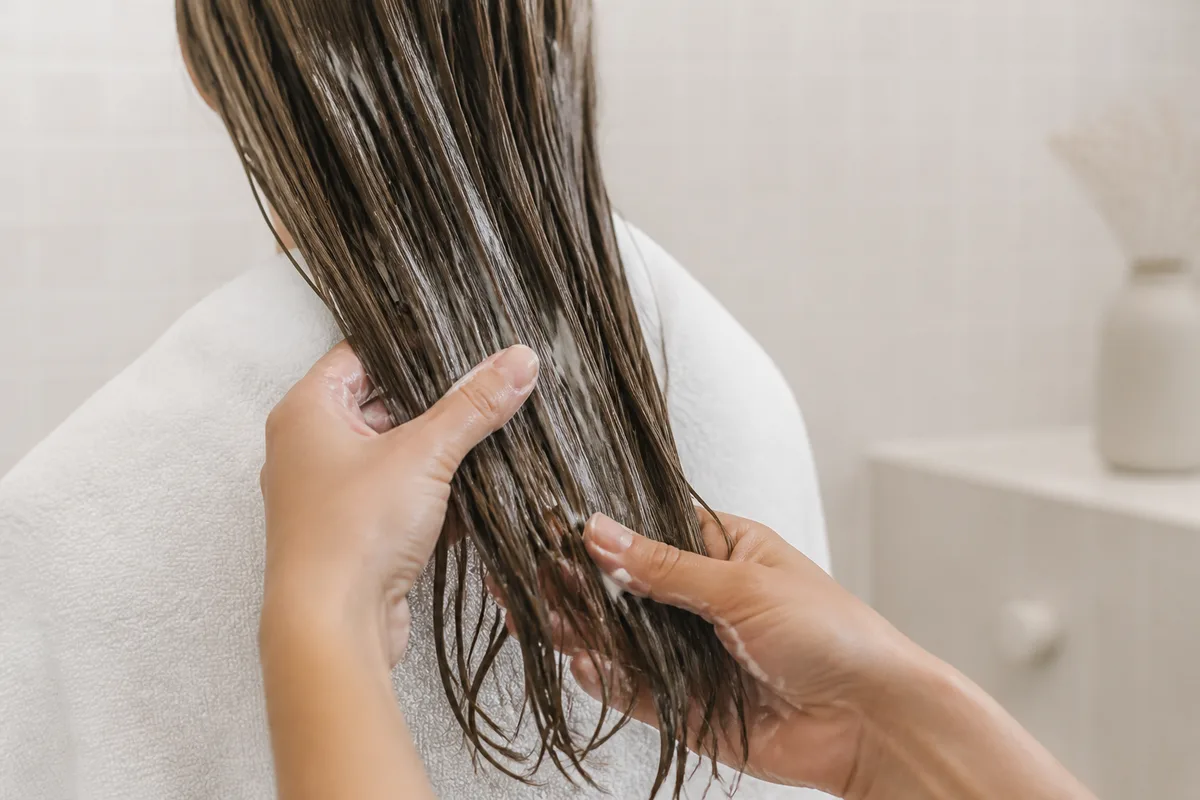 Cheveux longs enduits d&rsquo;un masque crémeux, mains en train de répartir le soin sur les longueurs